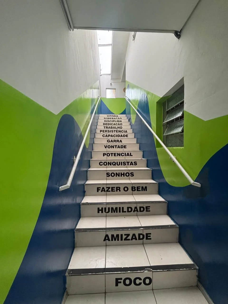 Escadaria azul e verde do Colégio JJR com valores escritos nos degraus, como amizade, humildade e foco